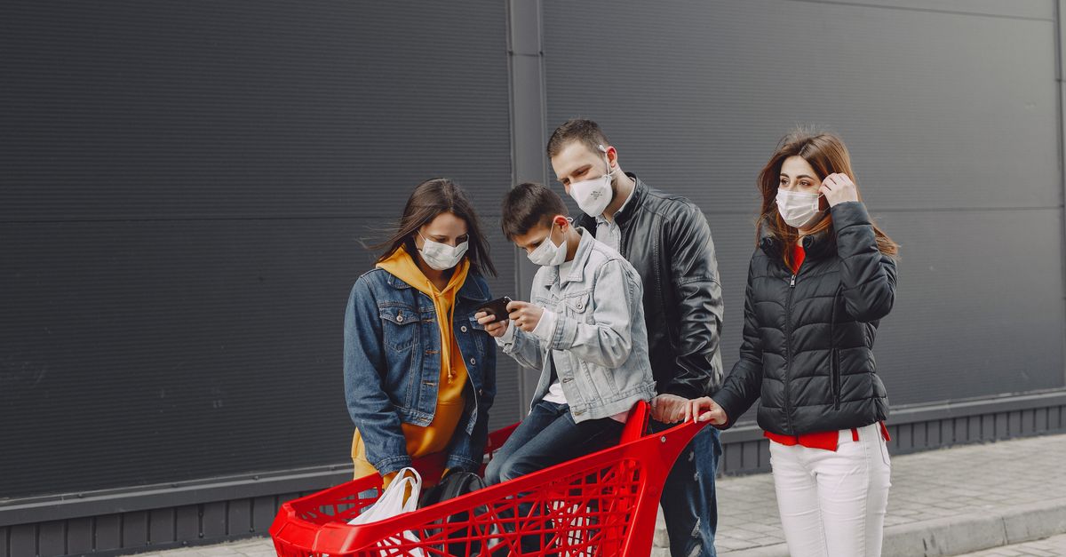 Happy family in casual clothes and medical masks standing on street after shopping while kid sitting in trolley and playing games on smartphone